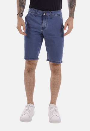 Uomo che indossa pantaloncini di jeans blu e sneakers bianche, in piedi con le mani rilassate ai lati davanti a uno sfondo semplice.