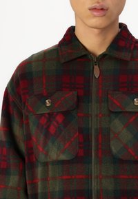 Rot-grünes karierter Fleece-Jacke mit Reißverschluss, zwei Brusttaschen und Kragen, aus weichem Material und stabilen Knöpfen gefertigt.