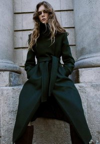 Mujer joven con cabello largo y ondulado que lleva un abrigo verde oscuro ceñido con cinturón, de pie frente a una pared de piedra con grandes columnas.