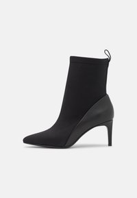 Svart spetstövlett med spetsig tå och smal hög klack, med en dragflik bak. Visar en slät, matt yta i tyg och läder.