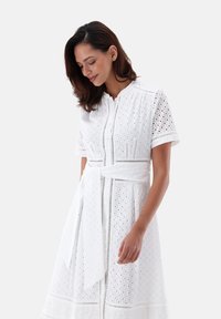 Weißes Baumwollkleid mit kurzen Ärmeln, Lochmuster, Knopfleiste und einem Gürtel, der in der Taille gebunden ist, mit einem ausladenden Rock.