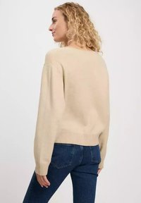 Beżowy sweter z dzianiny o luźnym kroju, z opadającymi ramionami, ściągaczem u dołu i długimi rękawami, w zestawieniu z ciemnoniebieskimi dżinsami.