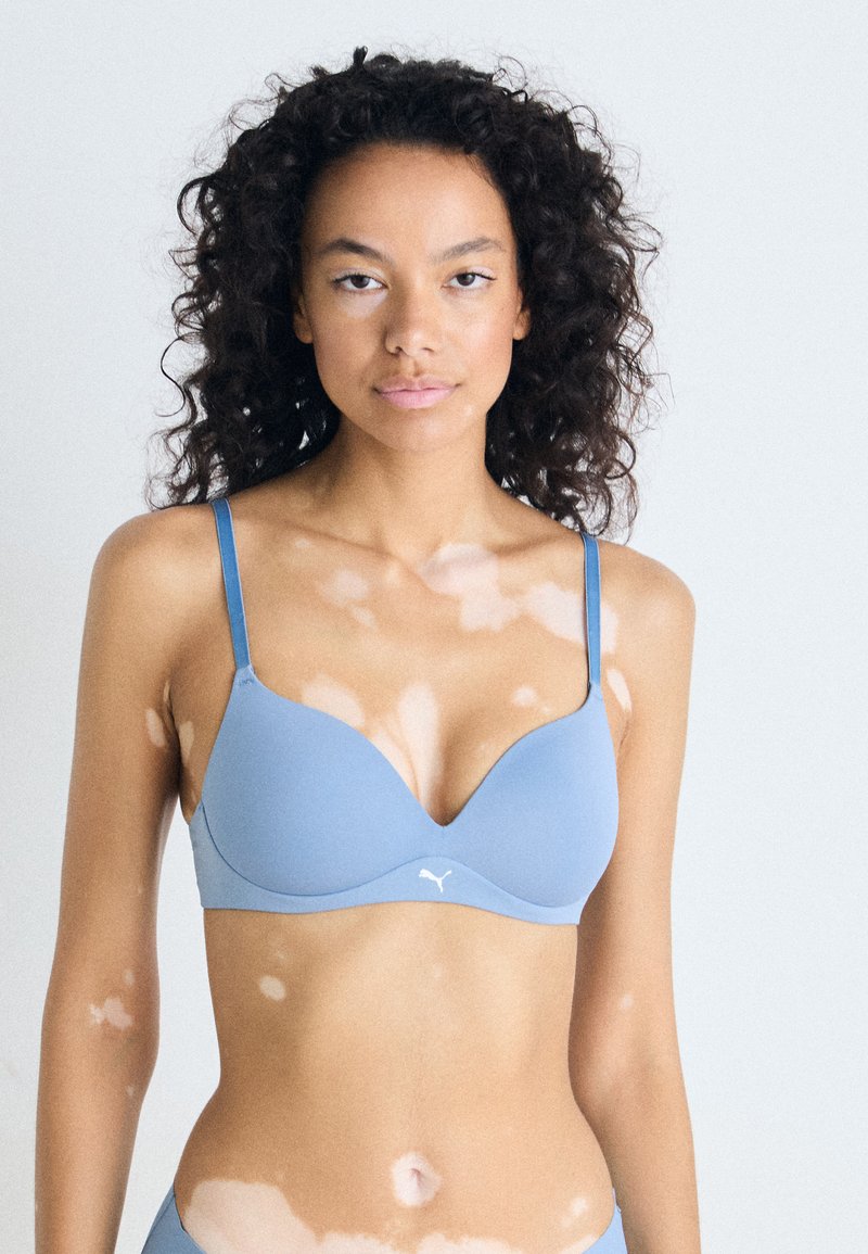Kvinne med krøllete hår som har på seg en lys blå bh, som viser vitiligo flekker i ansiktet, på armene og torsoen, står mot en ensfarget bakgrunn.