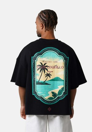 Man with braided hair wears black t-shirt with tropical beach graphic and text "Playa Luquillo" on back, paired with white pants.