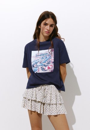 Young woman with long hair wearing a navy t-shirt with a beach umbrella print and a white patterned layered mini skirt.