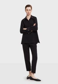 Musta ylisuuri paita, jossa on kaulus, pitkät hihat ja napit, yhdistettynä vartalonmyötäisiin mustiin housuihin ja mustiin kengissä. Silkkinen kangastunne.