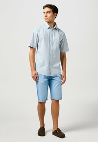 Camisa de rayas azul claro de manga corta con botones en la parte delantera, combinada con pantalones cortos de mezclilla claros. El modelo lleva zapatos marrones tipo slip-on.
