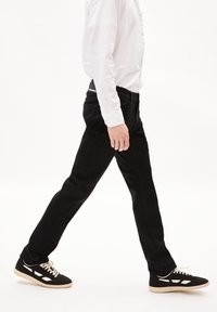 Jean noir avec une coupe droite, design classique à cinq poches, porté avec des baskets noires et blanches. La chemise est blanche avec des détails de boutons.