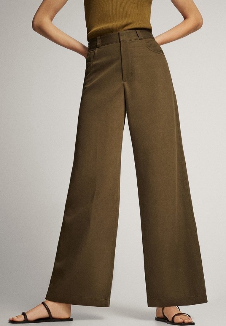 Femme portant un pantalon marron taille haute à jambes larges, associé à des sandales plates noires et un haut sans manches rentré.