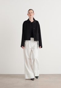 Zwarte cropped jack met ronde kraag, knoopsluiting; gecombineerd met losse witte broek en puntige grijze schoenen op een neutrale achtergrond.
