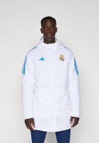 White insulated jacket with a hood, blue stripes, and Real Madrid logo on the chest. Features long sleeves and a straight cut.