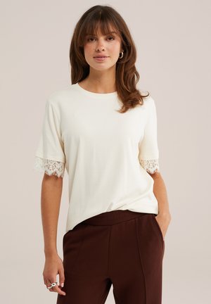 Femme aux cheveux bruns portant un haut blanc à manches courtes avec des détails en dentelle sur les manches et un pantalon marron foncé, debout, une main dans la poche.