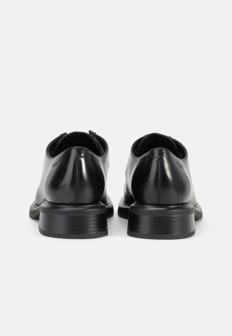 Par de zapatos de vestir de cuero negro con tacones apilados, vistos desde atrás sobre un fondo blanco.