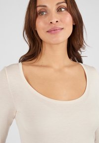 Lichtbeige langwerpige top met een ronde halslijn en een zachte, gladde textuur. Subtiele stiksels rond de halslijn verbeteren het ontwerp.