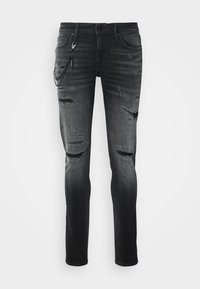 Jean slim noir déchiré avec des empiècements usés, un accessoire chaîne attaché à la poche avant gauche, et une fermeture à bouton et zip.