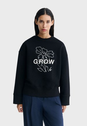 Jeune femme aux longs cheveux foncés porte un sweat-shirt noir avec un croquis de fleur blanche et le mot "GROW," assorti à un pantalon bleu foncé.