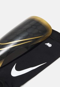 Zwarte voetbalschuinbescherming met een hexagonaal patroon, voorzien van een gladde, glanzende afwerking. Een opvallend wit Nike-logo en gouden accenten.