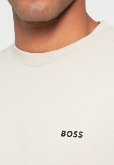 Sweat-shirt beige clair à col ras du cou avec col côtelé, arborant un logo "BOSS" noir sur le devant. Texture de tissu douce.