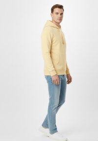 Sudadera amarilla de tela suave con un bolsillo frontal, combinada con vaqueros azul claro y zapatillas blancas, mostrada desde un perfil lateral.