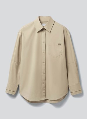Hosszú ujjú, bézs színű gombos ing gallérral és elülső mellzsebbel, világosszürke háttér előtt laposan elterítve.