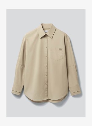 Hosszú ujjú, bézs színű gombos ing gallérral és elülső mellzsebbel, világosszürke háttér előtt laposan elterítve.