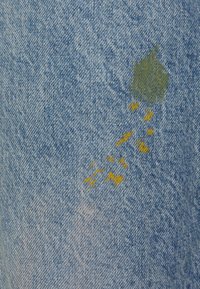 Tela de mezclilla de color azul claro, con marcas de pintura verde y amarilla cerca del centro, creando un patrón texturizado y multicolor.