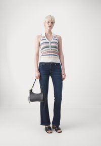 Top halterneck tricotado com riscas multicoloridas, combinado com jeans flare azul escuro. A modelo segura uma mala cinza texturizada, usando sandálias pretas.