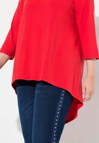 Maglietta rossa a maglia con maniche a tre quarti e orlo curvo, abbinata a jeans blu scuro con una fila di borchie argentate lungo il lato.