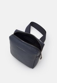 Borsa in pelle blu navy con superficie texture e bordi arrotondati. Presenta una chiusura con cerniera e una tracolla in tessuto. Logo Calvin Klein visibile.