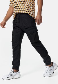 Sorte cargo-bukser med elastiske manchetter, flere sidelommer og en afslappet pasform, parret med hvide og grå sneakers. T-shirt med geometrisk mønster.