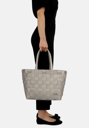 Gray woven tote bag with stitched texture, two braided handles, and silver hardware accents, held by a person in black attire.