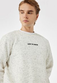Sudadera gris claro con un patrón texturizado de zigzag, que presenta texto bordado en negro "PERSEGUIR EL HORIZONTE" en el centro. Cuello redondo.