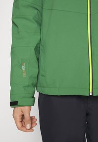 Persona che indossa una giacca softshell verde con zip e pantaloni neri, mano rilassata lungo il fianco, logo della giacca e fascetta sul polsino visibili.