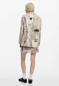 Beige blazer with a collage of prints and patterns, paired with a sheer beige skirt. Model wearing black sneakers and white socks.