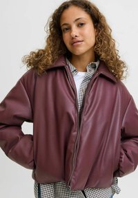 Bordowa skórzana kurtka z szerokim kołnierzem, zapinana na zamek, w luźnym kroju, noszona na czarno-białą koszulę w kratę. Gładka tekstura, minimalistyczny design.