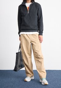 Mörkgrå sweatshirt med kvartdragkedja och framficka, beige vida byxor och vita sneakers med gröna detaljer. Svart axelväska i handen.