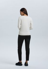 Lichtbeige lange mouw top met een gladde textuur, gecombineerd met zwarte slim-fit broek en zwarte sneakers met gouden accenten, gezien van achteren.