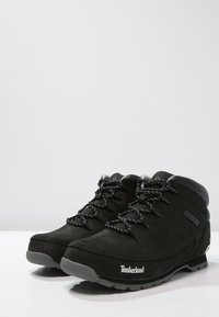 Botas de senderismo negras de ante con un acabado suave, que cuentan con suelas de goma gris y cordones negros con un patrón. Logo de Timberland visible.