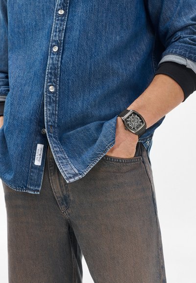 Jeanshemd in Mittelblau mit Knopfleiste und Manschettendetails, kombiniert mit dunkelbraunen Jeans. Schwarze rechteckige Uhr am Handgelenk.