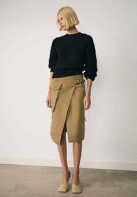 Maglione nero a coste con scollatura rotonda, abbinato a una gonna avvolgente color khaki con tasche laterali e dettaglio ad anello. Scarpe beige con tacco quadrato.