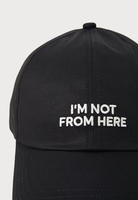 Svart baseball caps med flat skjerm, med hvit brodert tekst som leser "JEG ER IKKE FRA HER" foran. Glatt stoff tekstur.