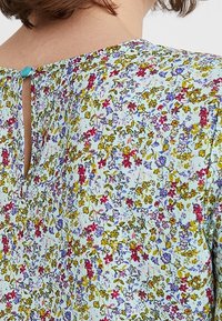 Květinová blůza se světle modrým pozadím, zdobená malými růžovými, žlutými a fialovými květinami. Výrazný klíčkový výřez vzadu a modrý knoflíkový detail.