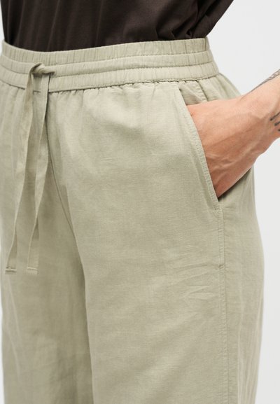 Μπεζ casual παντελόνι με ελαστική μέση και κορδόνι, χέρι στην πλαϊνή τσέπη, ορατό τατουάζ στον καρπό, φορεμένο με σκοτεινό τοπ.