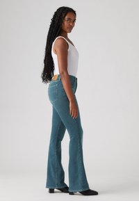 Jeans azuis à boca de sino com cintura média, cinco bolsos e um patch de couro castanho na parte de trás da cintura. O modelo veste uma t-shirt branca.