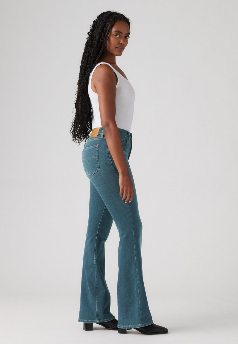 Jeans a palazzo blu con vita media, cinque tasche e una patch in pelle marrone sulla parte posteriore della vita. Il modello indossa una canotta bianca.