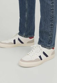 Witte sneakers met marineblauwe strepen en lichtgrijze accenten, voorzien van een gestructureerde rubberen zool, ronde neus en vetersluiting, gecombineerd met blauwe jeans.