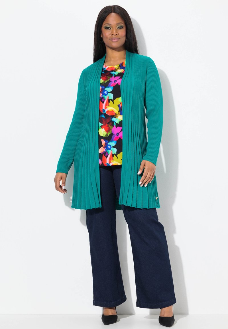 Teal, ribbad cardigan med öppen framsida och långa ärmar, kombinerad med en färgglad blommig topp och mörka jeans, kompletterad med svarta skor.