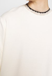 Felpa color crema con un collo a strisce nere e beige testurizzato. Il tessuto sembra morbido con una finitura liscia.