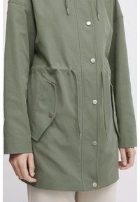 Veste légère et verte avec une taille à cordon de serrage, fermeture à pression et deux poches latérales. Présente une texture lisse et des accents métalliques.