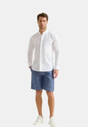 Camicia bianca a maniche lunghe con collo alla cinese, abbinata a pantaloni corti blu a fantasia e sneaker bianche, in piedi su uno sfondo neutro.